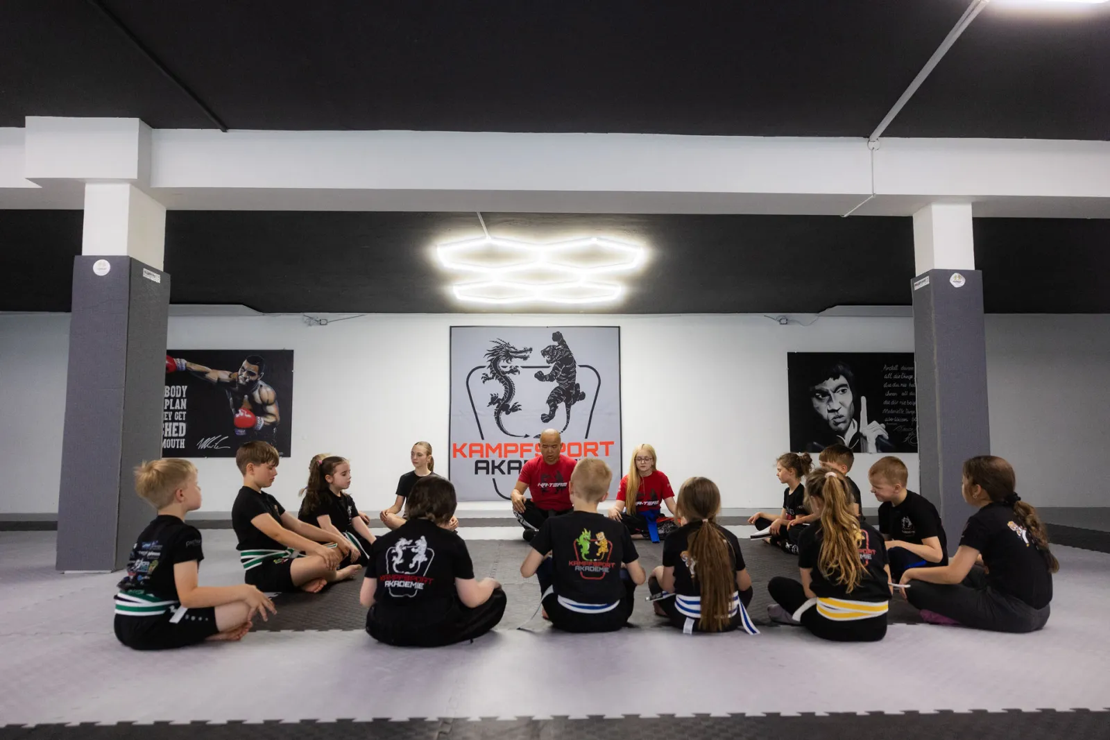 Sitzkreis zum Unterrichtsbeginn vor der Logo-Wand der Kampfsportakademie Mengen